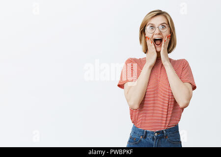 Heureux heureux et joyeux ventilateur femelle aux cheveux blonds dans les verres, des cris de joie et d'excitation, tenant les mains sur les joues d'être impressionné et submergés d'émotions en voyant célèbre rockstar Banque D'Images