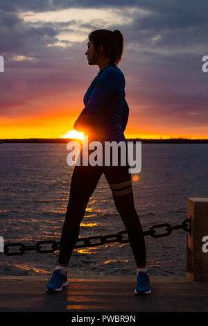 Une jolie femme en vêtements de sport Le sport exercices dans la nature contre le coucher du soleil et le fleuve Amour, aime la gymnastique, pétrit ses jambes. Jeune fille active engagée dans le sport, mène une vie saine. Banque D'Images