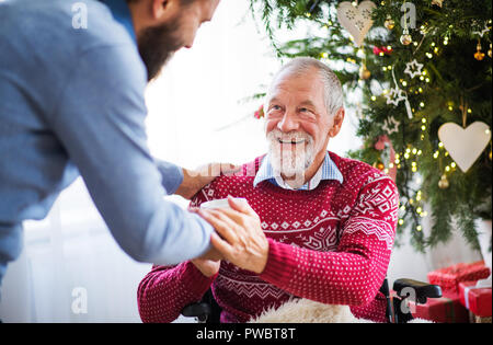 Un homme hipster méconnaissable en donnant une boisson dans une tasse à ses hauts père en fauteuil roulant à l'époque de Noël. Banque D'Images