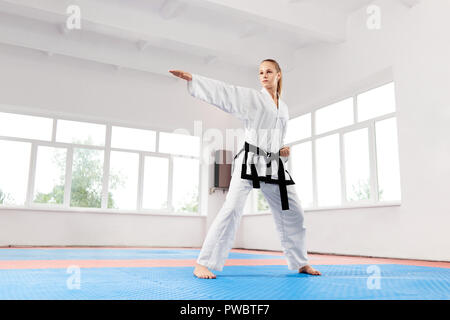 Karaté sportif femme en kimono blanc avec ceinture noire karate contre grande fenêtre à lutte de classe. Chasse femelle avec les yeux bleus et les cheveux tressés prêts à attaquer son adversaire. Banque D'Images