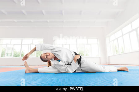 Young Girl wearing active en kimono blanc avec ceinture noire, faire de l'exercice sur la flexibilité. Femme sportive sérieusement de souplesse jusqu'à l'étage avant l'entraînement à la lumière de sport. Concept de vie sain. Banque D'Images