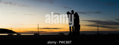 Vue arrière de la silhouette du famille en plein air à étreindre au coucher du soleil avec la mer, la montagne et voiliers derrière dans une large vue panoramique contre e Banque D'Images