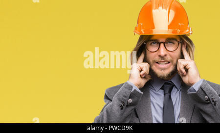 Beau jeune homme architecturaux aux cheveux longs portant un casque de sécurité sur fond isolé couvrant les oreilles avec les doigts avec l'expression ennuyé pour la n Banque D'Images