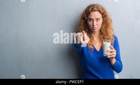 Jeune rousse femme à boire le verre de lait avec le doigt pointant à l'appareil photo et à vous, signe de main, geste positif et confiant de l'avant Banque D'Images