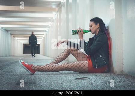 Une jolie jeune fille assise sur le trottoir et de boire une bouteille de whisky en séparation avec un être cher. girl a rompu avec son petit ami Banque D'Images