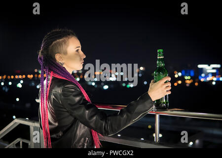 Portrait de femme seule boire de l'alcool à partir de la bouteille à l'autoroute. Une jeune fille ivre avec une bouteille de whisky se dresse sur le toit de la maison et regarder Banque D'Images