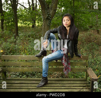 Jeune femme en veste en cuir noire en bois/forêt assis sur le vieux banc en bois recouverte de mousse en milieu urbain. vêtements et jeans. Banque D'Images