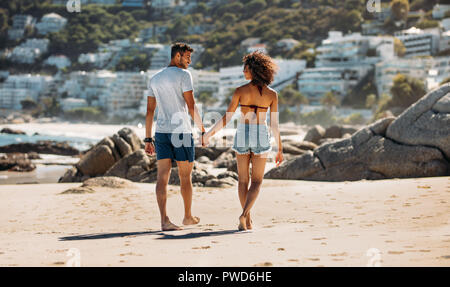 Couple de touristes en vacances à la plage sur la marche vers l'autre. Vue arrière d'un couple en train de marcher sur une plage rocheuse, main dans la main sur une journée ensoleillée. Banque D'Images