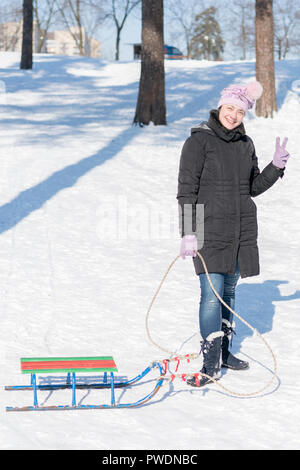 Une femme dans un manteau d'hiver noir avec traîneaux dans un parc couvert de neige ou la forêt. photo verticale. Banque D'Images