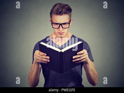 Jeune homme dans les verres à confondre tout en essayant de lire smart livre sur fond gris Banque D'Images