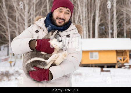 Jusqu'à la taille de l'homme asiatique moderne portrait smiling at camera while holding mignon chiot Husky à l'extérieur en hiver, copy space Banque D'Images