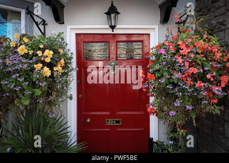 Deux grands paniers de fleurs multicolores pendent de chaque côté d'une porte avant rouge vif sur une maison blanche à Thornbury, Royaume-Uni, Britain in Bloom contest. Banque D'Images