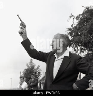 1967, l'homme à l'aide d'un fusil ou pistolet starter démarreur extérieur, à une fête du village, d'annoncer le début de la lutte à la corde, concours d'argent, England, UK. Un starter gun est un pistolet vide et est tiré pour lancer des concours sportifs, notamment les courses d'athlétisme. Banque D'Images