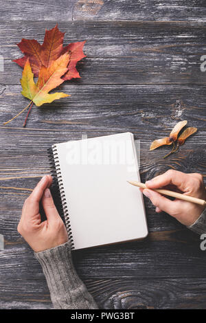 Avec la maquette papier blanc décoré d'ordinateur portable les feuilles d'automne sur la table en bois. Woman's hand writing dans le bloc-notes. Vue de dessus, à la verticale. Banque D'Images