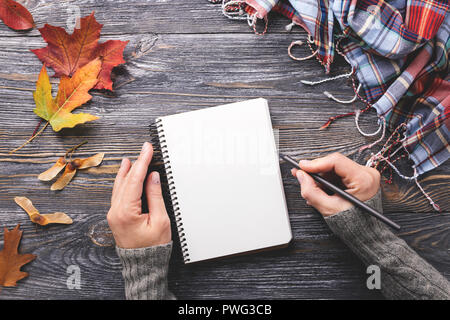 Avec la maquette papier blanc décoré d'ordinateur portable et les feuilles d'automne foulard à damiers sur table en bois. Woman's hand writing dans le bloc-notes. Vue d'en haut. Banque D'Images