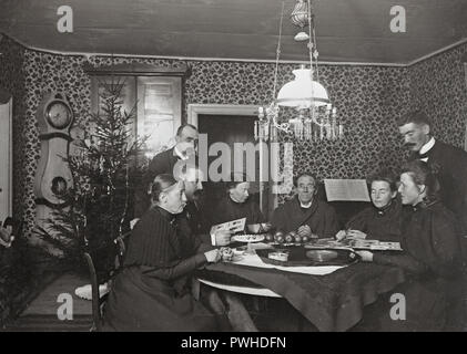 Noël au tournant du siècle, 1800-1900. Un prêtre a une place centrale à la table où la famille est assis. C'est vacances de Noël et ils profitent de la soirée à la recherche au sein du cabinet photos de famille et de manger des pommes de noël. Un arbre de Noël est debout dans le coin. La Suède 1800 REF 379A-13 Banque D'Images