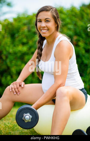 Portrait of smiling femelle enceinte l'entraînement avec haltères en position assise sur le yoga ball pour renforcer en armes park Banque D'Images