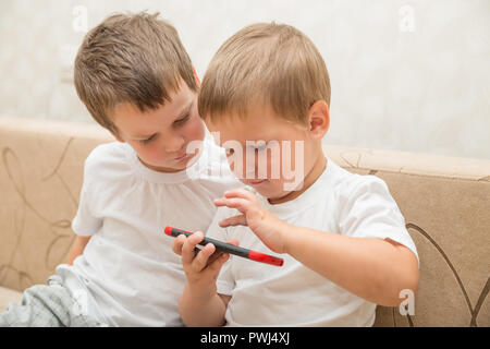 Deux petits garçons en blanc T-shirts sont assis sur le canapé à la maison. Un garçon joue à des jeux sur smartphone, deuxième on regarde Banque D'Images