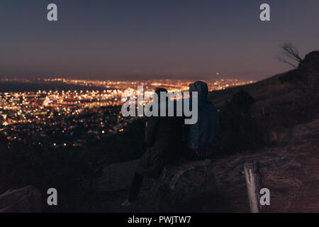 Couple watching la ville illuminée assis au sommet d'une colline tard dans la soirée. Vue arrière du couple sur une colline et bénéficiant d'une conversation Banque D'Images