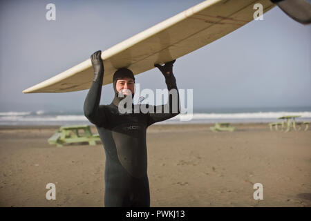 Portrait of a smiling young man wearing une combinaison isothermique et transportant un surf sur sa tête sur une plage de sable.