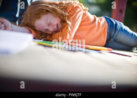 Jeune fille riant tout en étant allongé sur le ventre à côté d'un livre de coloriage et des crayons à l'extérieur dans le soleil. Banque D'Images