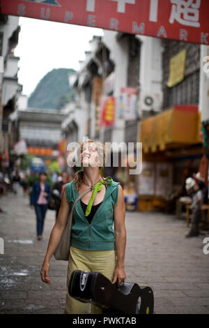 Young Woman smiling en marchant le long d'une rue portant un étui à guitare. Banque D'Images