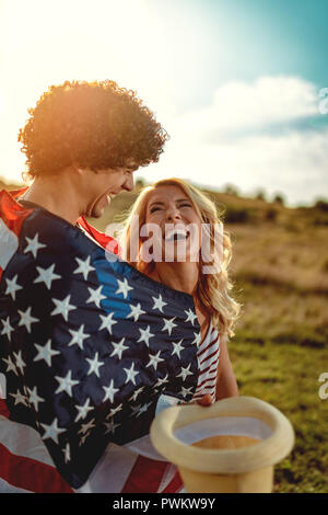 Happy young couple bénéficie d'une journée ensoleillée dans la nature. Ils êtes serrant les uns les autres, enveloppés dans le drapeau américain. Banque D'Images