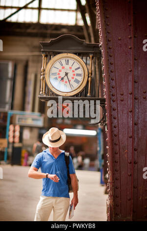 Jeune homme vérifie sa montre et est titulaire d'une cigarette et d'un livre comme il se tient sous une horloge dans une gare. Banque D'Images