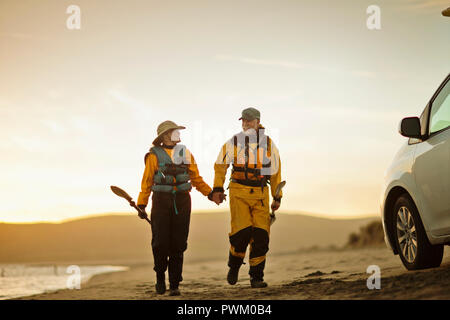 Happy senior couple marche main dans la main tout en portant des pagaies le long d'une plage de sable. Banque D'Images