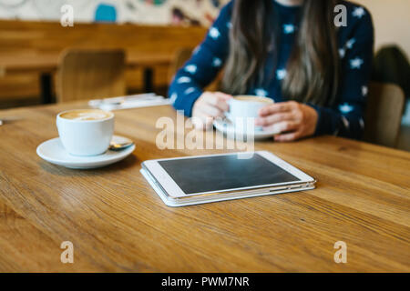 Cappuccino dans une tasse sur la table à côté de la tablette. La fille est de boire du café dans l'arrière-plan. Se détendre ou à une réunion dans un café. La fille est en attente de quelqu'un d'autre. Banque D'Images