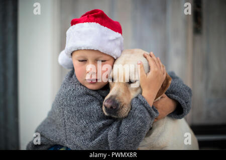 Young boy wearing a santa hat ferme les yeux alors qu'il embrasse son chien et les rêves de Noël. Banque D'Images