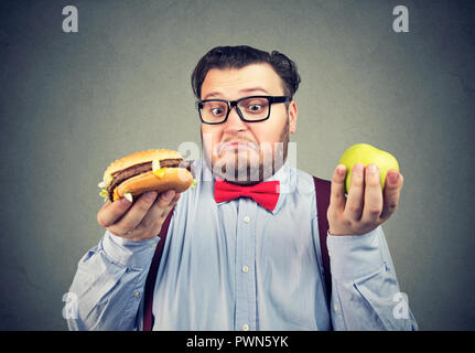 Jeune homme grassouillet en nœud papillon et verres holding apple et burger à la confondre avec choix Banque D'Images