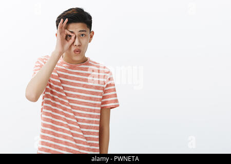 Zéro soucis. Portrait de jeune garçon asiatique élégant T-shirt à rayures en faisant bon geste sur eye peeking through doigts lèvres pliage surpris et intrigué posing against white background Banque D'Images