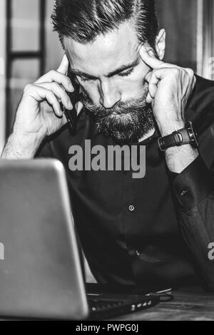 Jeune homme triste barbu est assis à table, couvrant son visage avec sa main et on cell phone. Le bureau est l'ordinateur portable, tablettes. Man Banque D'Images