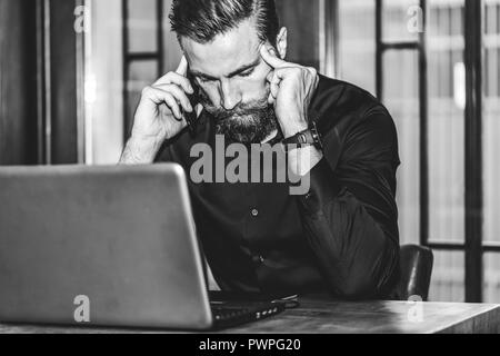 Jeune homme triste barbu est assis à table, couvrant son visage avec sa main et on cell phone. Le bureau est l'ordinateur portable, tablettes. Man Banque D'Images