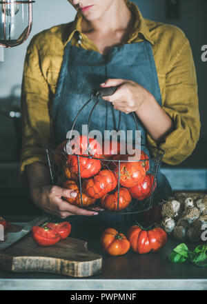 Femme en tablier gris holding basket with heirloom tomatoes et la cuisson de la sauce tomate, tomates en conserve ou de pâtes au basilic et l'ail à la cuisine Banque D'Images