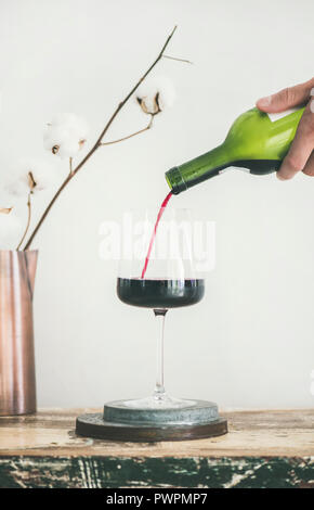 Verseur à vin rouge de la bouteille dans la main de l'homme en verre sur un comptoir de cuisine rustique, fond blanc derrière, composition verticale. Wine Shop, wi Banque D'Images