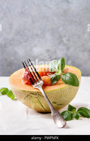 Melon et jambon cru ou salade de prosciutto servi dans la moitié de melon cantaloup, décoré par le basilic frais debout sur nappe blanche avec une fourchette. Banque D'Images