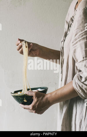 Femme en robe blanche tenant en mains bol de soupe de nouilles de riz asiatiques et prendre les nouilles par baguettes. Mur gris en arrière-plan. Dîner de style asiatique. Banque D'Images