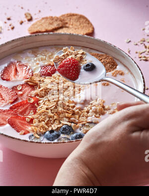 Prend la main de femme avec une cuillère de yogourt biologique , smoothie fraises, myrtilles, flocons d'avoine, de noix - dessert de fruits sur fond rose. Banque D'Images