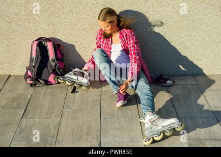 Teen girl supprime les espadrilles et les vêtements des patins à l'extérieur. Siège au mur, à côté de son sac à dos. Banque D'Images