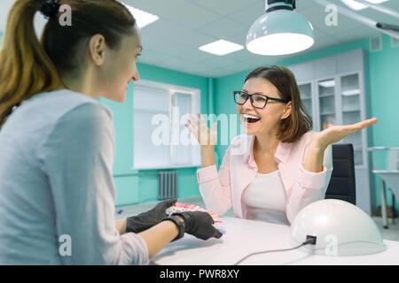 Jeune fille esthéticienne manucure parle à une cliente, manucure, ne choisit la couleur de vernis à ongles, communique et sourit. Banque D'Images