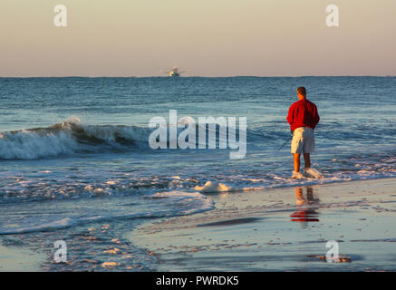 Un homme pêche ocean surf au petit matin avec un bateau de crevettes à l'arrière-plan. Banque D'Images