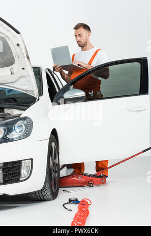 Beau mécanicien d'automobile en uniforme orange holding laptop près de voiture cassée isolated on white Banque D'Images