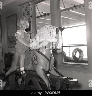 Années 1950, historique, sur un bateau, dans la pépinière ou de jeu, une jeune fille est assise jouant sur un cheval à bascule en bois par la fenêtre. Banque D'Images