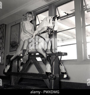 Années 1950, historique, sur un bateau, dans la pépinière ou de jeu, une jeune fille est assise avec ses pieds dans le jeu stirups ou équitation sur un cheval à bascule en bois par la fenêtre. Banque D'Images