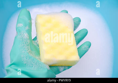 Une main dans un gant turquoise est titulaire d'une éponge à vaisselle dans une abondante mousse sur un fond bleu. Close-up Banque D'Images