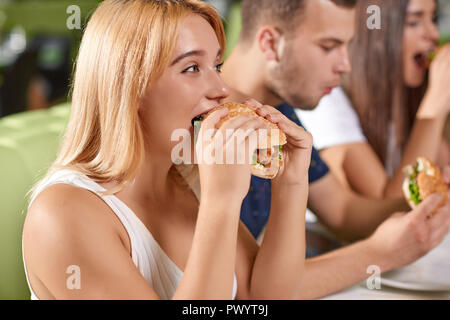 Vue latérale du jeune blonde affamé mordant burger juteux à nice cafe. Jolie jeune fille assise dans un café avec des amis et profiter de délicieux repas rapides. Concept de manger et de se détendre. Banque D'Images