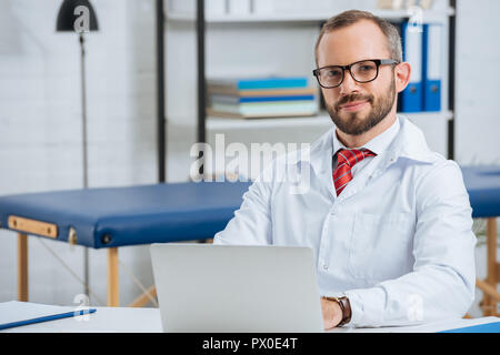 Portrait of smiling male chiropraticien en robe blanche sur le lieu de travail avec un ordinateur portable à l'hôpital Banque D'Images