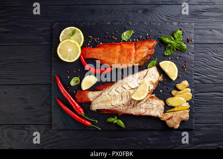 Filet de vivaneau rouge fumé à chaud sur une plaque en pierre noire avec des épices, des herbes et des tranches de citrons, vue de dessus, close-up Banque D'Images